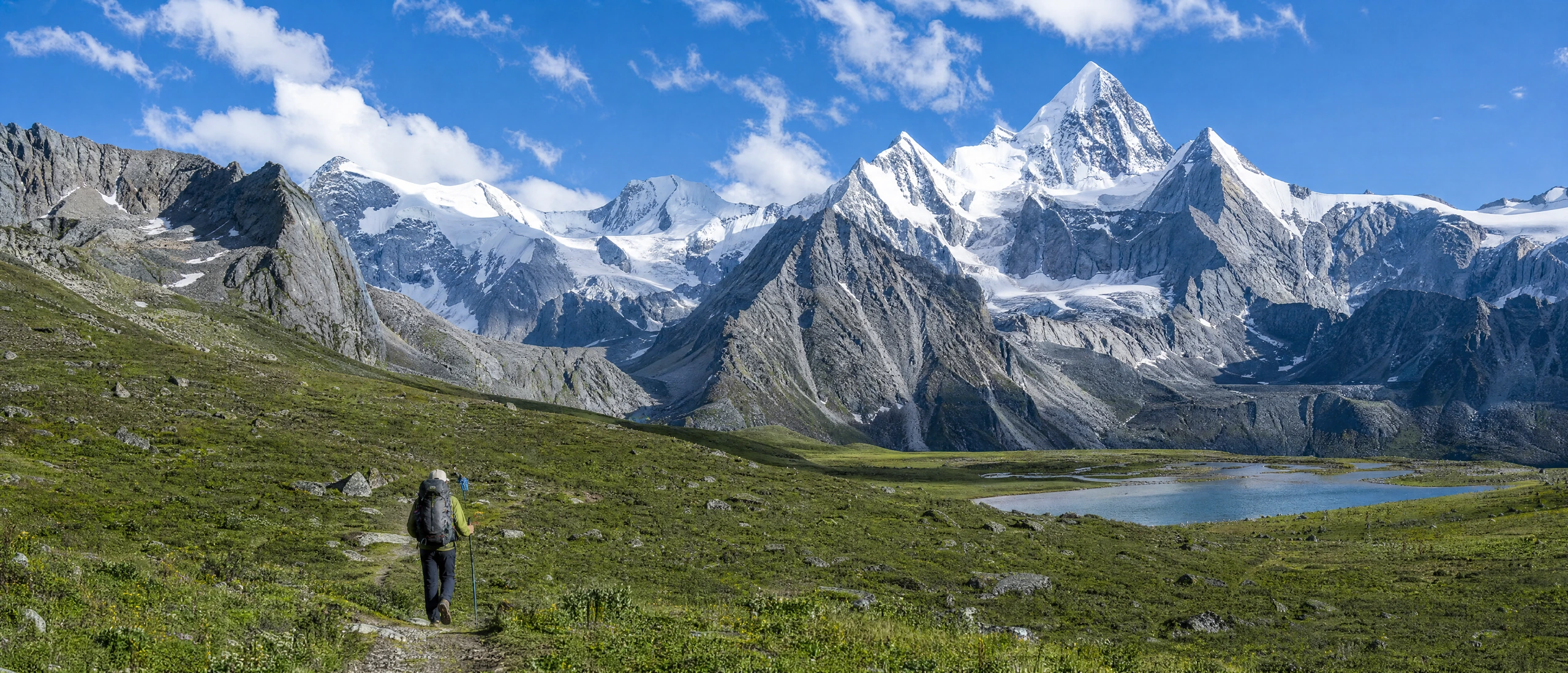 A hiker crossing a meadow beside a lake beneath snowy peaks